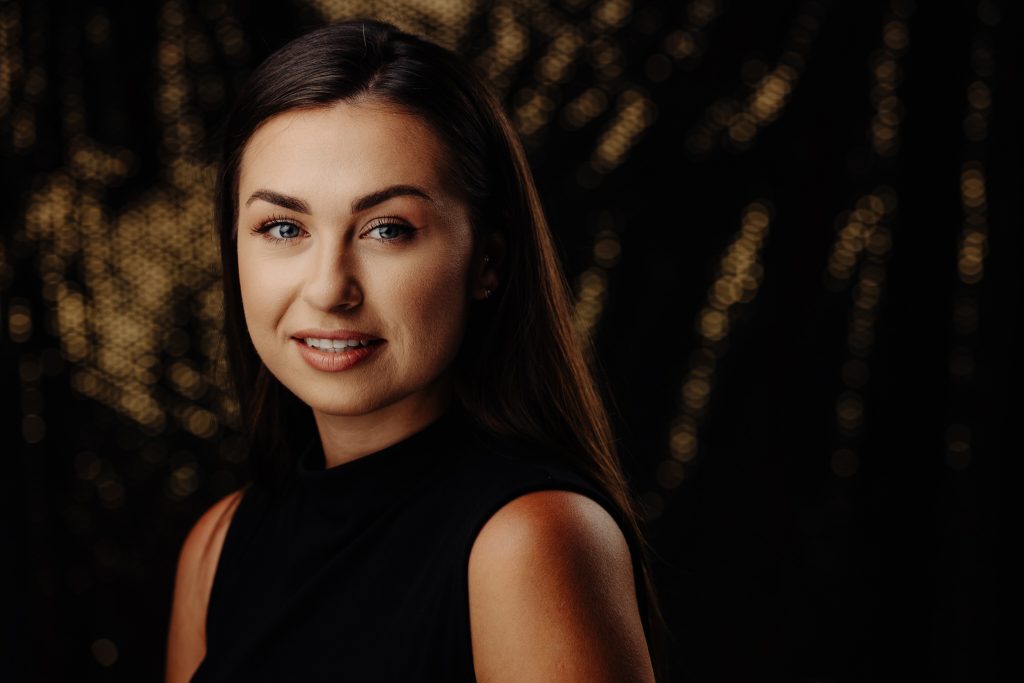 Cinematic Headshot: uśmiechnięta kobieta w czarnej bluzce, ciemne tło z ciepłym bokeh